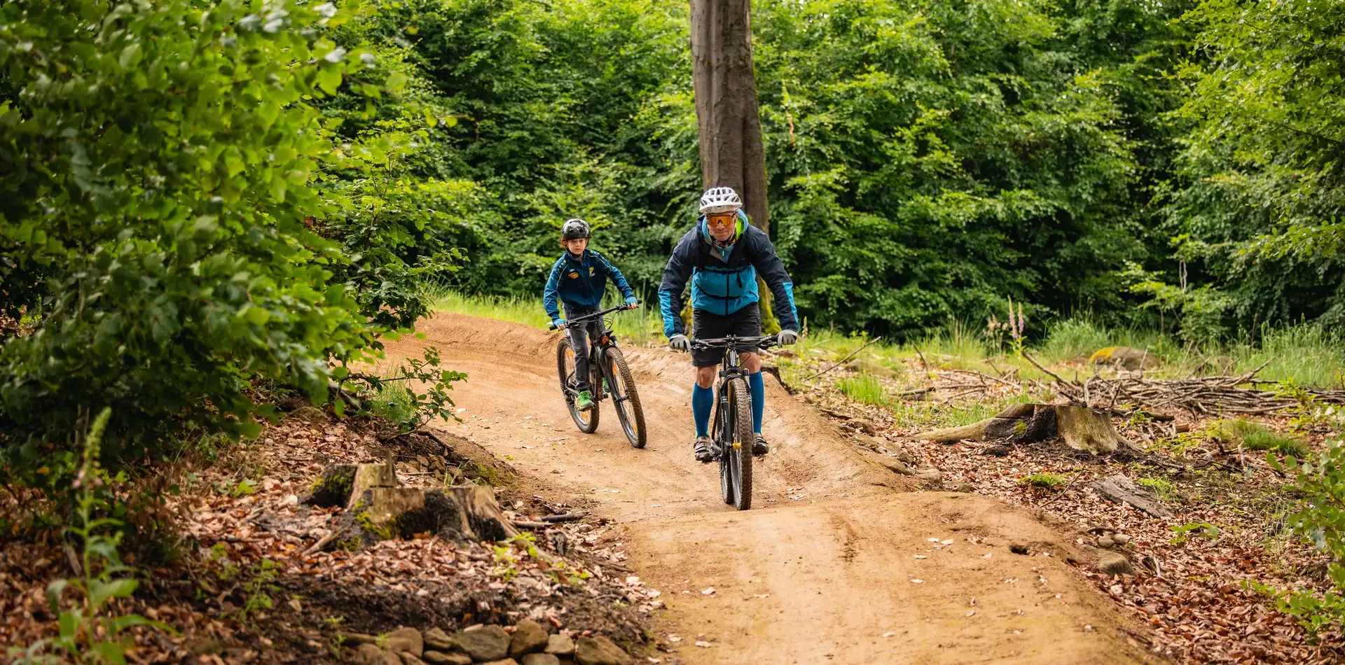 Zwei Mountainbiker fahren auf einem geschwungenen Trail durch den Wald.